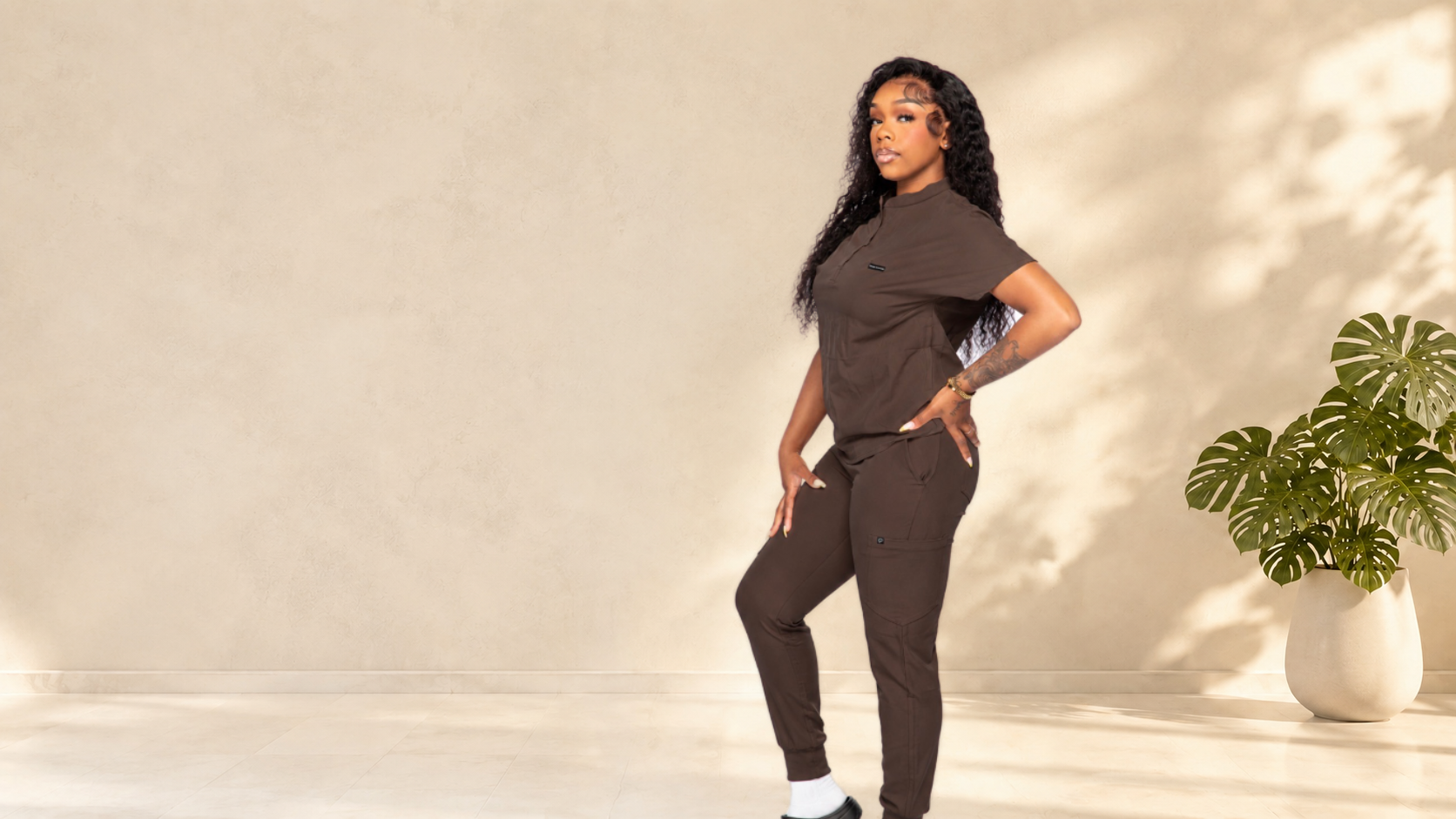 A woman modeling a matching dark brown short-sleeve top and jogger pants set in a minimalist studio with a beige wall, soft shadows, and a potted green plant on the right.